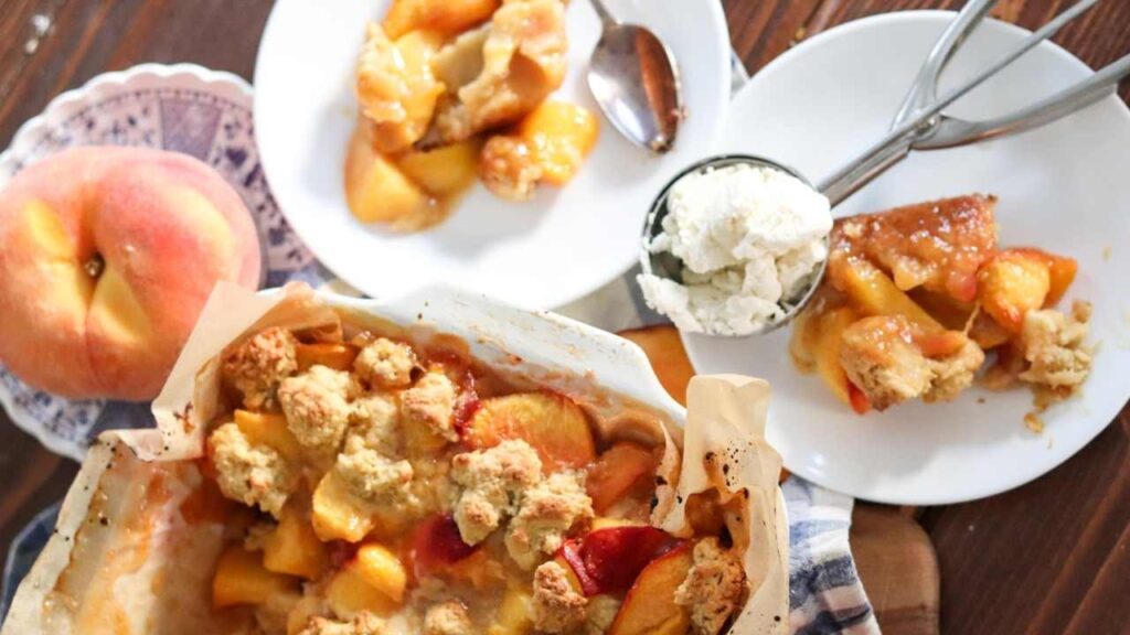 fresh peach cobbler with dishes next to pan of individual servings and a spoonful of ice cream.