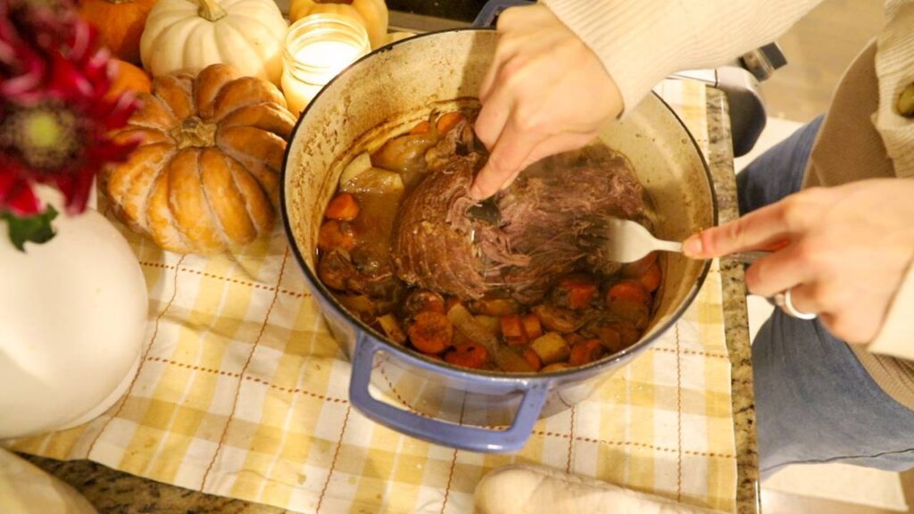 Cooked beef roast being shredded inside a Dutch oven with vegetables.