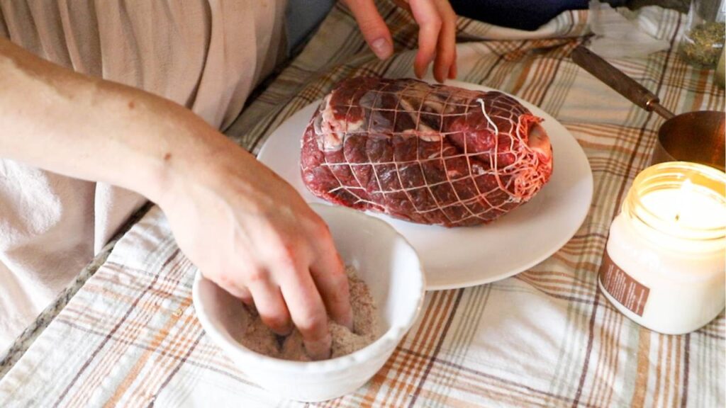 Hands seasoning a tied beef roast on a plate next to a bowl of spices.