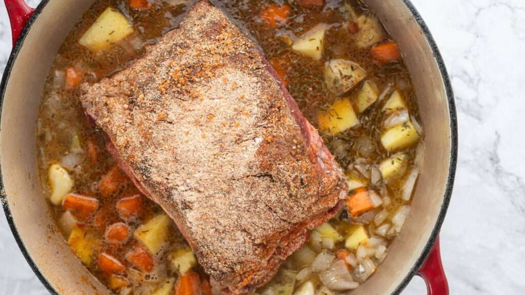 Seasoned beef roast resting in broth with carrots, onions, and potatoes in a Dutch oven.