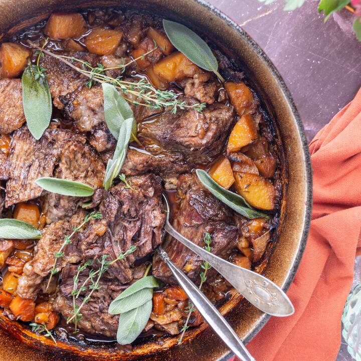 Dutch oven filled with apple cider pot roast, carrots, and potatoes topped with fresh herbs.