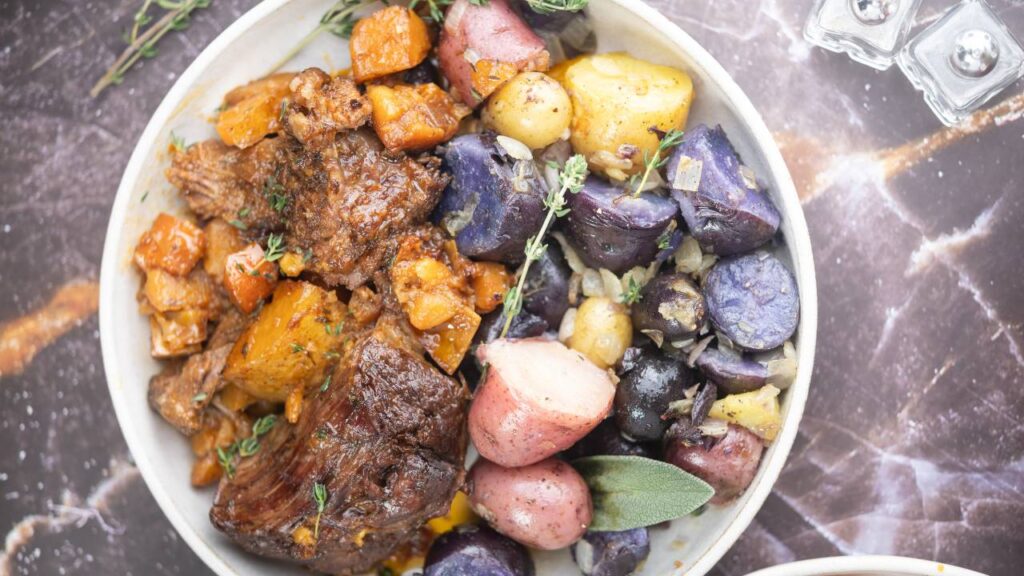 Dutch oven filled with apple cider pot roast, carrots, and potatoes topped with fresh herbs.