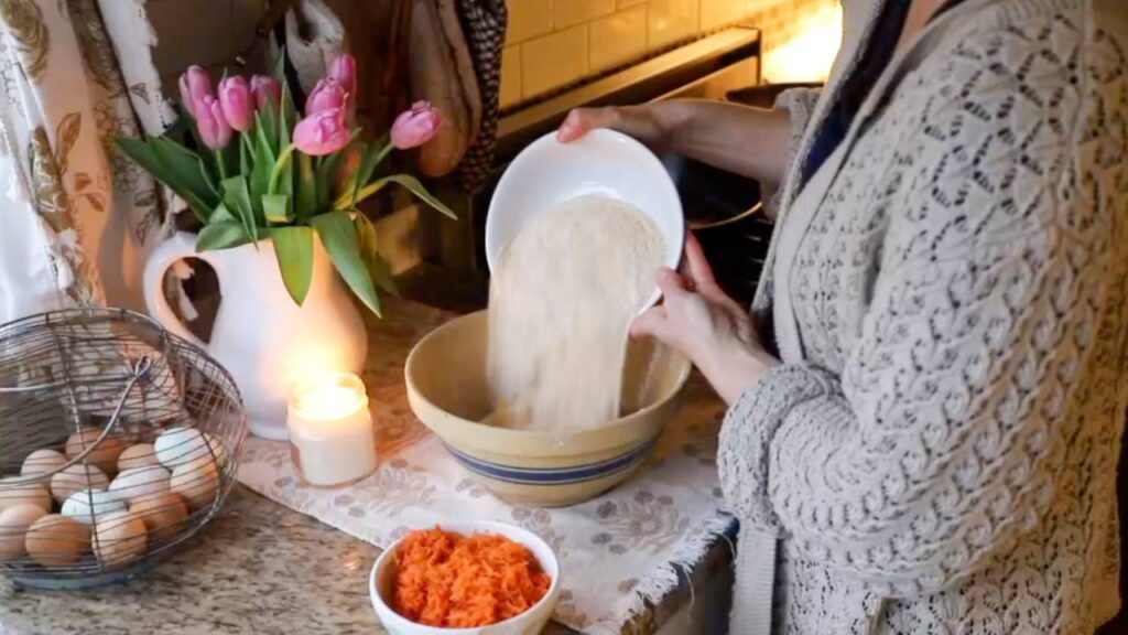 Pouring whole grain flour into a mixing bowl with grated carrots nearby.