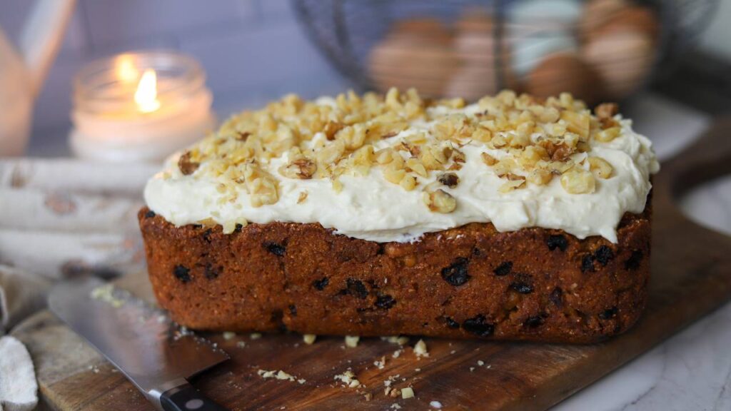 Side view of frosted carrot cake loaf with walnuts on a cutting board.